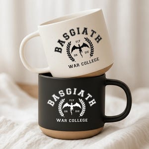 May include: Two ceramic mugs stacked vertically. The top mug is cream with black speckles and black text reading BASGIATH WAR COLLEGE with a dragon graphic. The bottom mug is matte black with white text and a matching dragon graphic design.