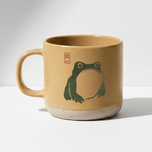 Könnte beinhalten: Eine beige Keramiktasse mit einer grünen Froschillustration und einem kleinen roten Quadrat mit japanischem Text. Die Tasse hat einen abgerundeten Henkel und einen gesprenkelten, cremefarbenen Sockel. Das Froschdesign ist eine einfache, stilisierte Zeichnung.