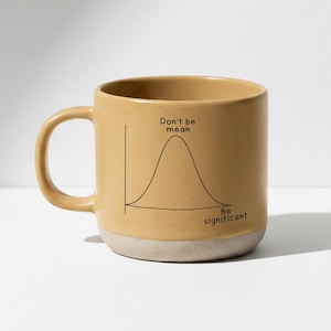 Op de afbeelding: Een bruin keramische koffiemok met een witte basis. De mok heeft een zwarte grafiek met de woorden "Don't be mean" en "Be significant". De mok heeft een gebogen handvat.
