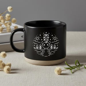 Peut inclure: Mug en c&eacute;ramique noire avec une base beige. Le mug pr&eacute;sente un dessin au trait blanc d'un personnage avec une &eacute;toile au-dessus de sa t&ecirc;te, entour&eacute; de fleurs, d'&eacute;toiles et de nuages. La tasse a une anse incurv&eacute;e.