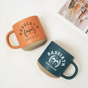 May include: Two ceramic mugs with handles, one orange and one dark blue. Both feature a circular emblem with a dragon silhouette, laurel branches, and the text Basgiath War College Fly or Die printed in a contrasting color on the front surface.
