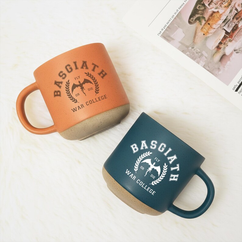 May include: Two ceramic mugs with handles, one orange and one dark blue. Both feature a circular emblem with a dragon silhouette, laurel branches, and the text Basgiath War College Fly or Die printed in a contrasting color on the front surface.