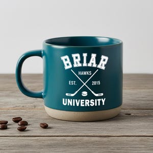 Puede incluir: Taza de cerámica color turquesa con el texto "Briar University" en blanco. La taza presenta palos de hockey cruzados y un gráfico de disco de hockey. La taza tiene una base beige y un asa grande. Granos de café esparcidos sobre la superficie de madera.