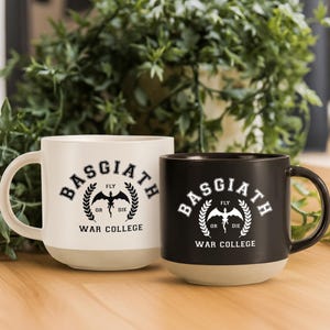 May include: Two ceramic mugs, one white and one black, each featuring a graphic print that reads Basgiath War College with a central dragon silhouette and the text Fly or Die. Both mugs have a smooth handle and a contrasting light beige base.