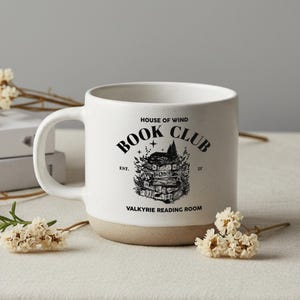 May include: White ceramic mug with a tan base and a large handle. The mug features a black graphic of a stack of books with a house on top, and the words "HOUSE OF WIND BOOK CLUB VALKYRIE READING ROOM" in black.