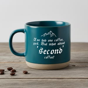 May include: A teal ceramic coffee mug with a white mountain graphic and the text "I've had one coffee, yes. But what about 'Second' coffee?". The mug features a smooth teal upper section and a speckled, unglazed beige base with a rounded handle.