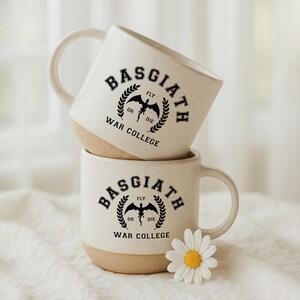 May include: Two stacked ceramic mugs with a speckled cream finish and a tan base. The mugs feature a black graphic with the text Basgiath War College, a dragon silhouette, and the words Fly or Die surrounded by a laurel wreath design.