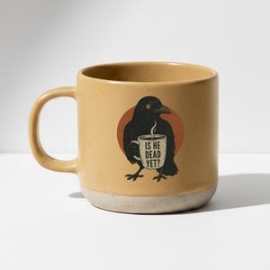 Peut inclure: Mug en céramique beige avec une illustration d'un corbeau noir tenant une tasse à café. La tasse à café porte l'inscription "IS HE DEAD YET ?". La tasse a une forme arrondie avec une anse et une base mouchetée blanc cassé.
