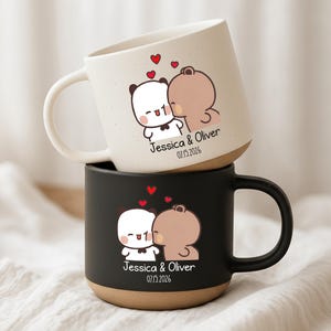 May include: Two ceramic mugs, one white and one black, stacked. Each mug features a cartoon illustration of two hugging figures with hearts above them. The mugs are personalized with the names "Jessica & Oliver" and the date "07.15.2026".