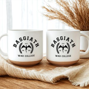 May include: Two white ceramic mugs with a natural clay-colored base. Each mug features a black graphic with the text BASGIATH WAR COLLEGE, a central dragon silhouette, and the words FLY OR DIE surrounded by a laurel wreath design on the front.