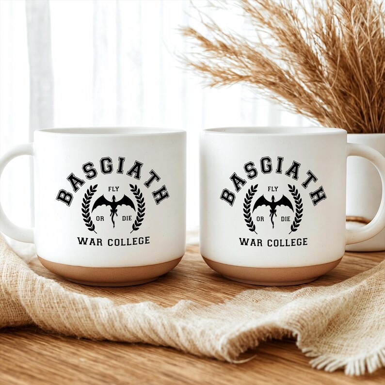 May include: Two white ceramic mugs with a natural clay-colored base. Each mug features a black graphic with the text BASGIATH WAR COLLEGE, a central dragon silhouette, and the words FLY OR DIE surrounded by a laurel wreath design on the front.