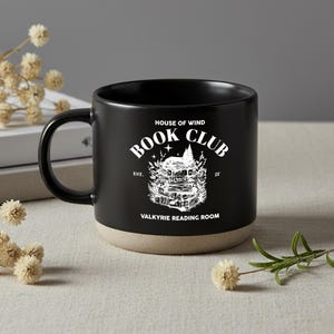 May include: Black ceramic mug with a beige base and a black handle. The mug features the text "HOUSE OF WIND BOOK CLUB" in white, along with an illustration of a stack of books. The words "VALKYRIE READING ROOM" are also printed on the mug.