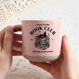 May include: A pink ceramic mug with the text "BOOK CLUB" and a graphic of stacked books. The mug also features the text "HOUSE OF WIND", "EST. 21'", and "VALKYRIE READING ROOM". The mug has a light pink handle and a gray base.