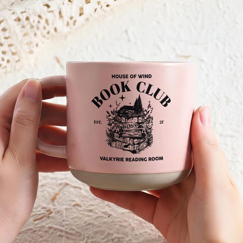 May include: A pink ceramic mug with the text "BOOK CLUB" and a graphic of stacked books. The mug also features the text "HOUSE OF WIND", "EST. 21'", and "VALKYRIE READING ROOM". The mug has a light pink handle and a gray base.