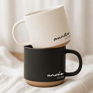 Può includere: Due tazze in ceramica impilate. La tazza superiore è bianca con macchie nere e la parola "auntie" e "EST. 2026" stampate sopra. La tazza inferiore è nera con la parola "uncle" e "EST. 2026" stampate sopra. Entrambe le tazze hanno una base marrone.