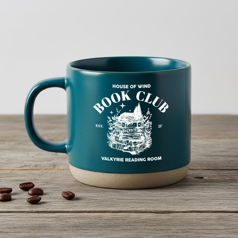 May include: Teal ceramic mug with a beige base and a large handle. The mug features the text "HOUSE OF WIND BOOK CLUB" and "VALKYRIE READING ROOM" in white, along with a detailed illustration of a stack of books. Coffee beans are scattered on the wooden surface.