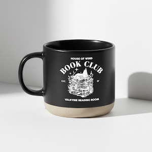 May include: Black ceramic mug with a beige base and a black handle. The mug features white text and a graphic that reads "House of Wind Book Club Valkyrie Reading Room."