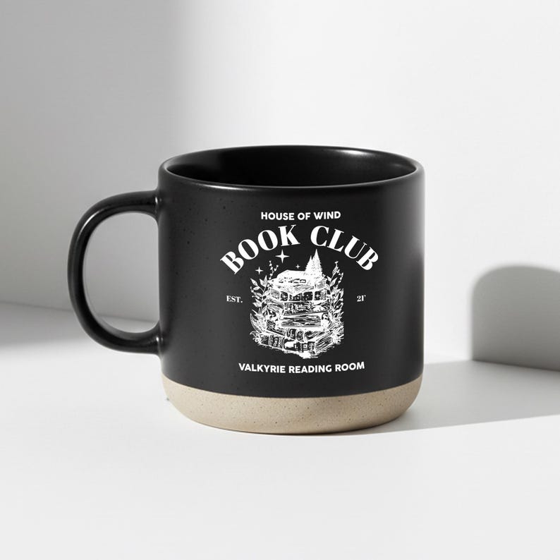 May include: Black ceramic mug with a beige base and a black handle. The mug features white text and a graphic that reads "House of Wind Book Club Valkyrie Reading Room."
