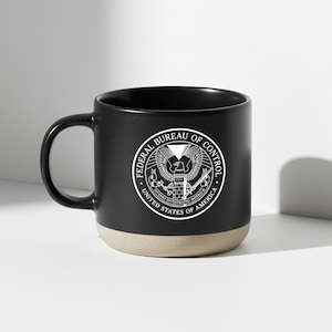 Könnte beinhalten: Eine schwarze Keramik-Tasse mit beigem Boden. Die Tasse hat ein weißes kreisförmiges Emblem mit dem Text "FEDERAL BUREAU OF CONTROL" und "UNITED STATES OF AMERICA". Die Tasse hat einen gebogenen Griff.