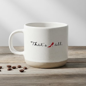 May include: White ceramic coffee mug with a speckled finish and a tan base. The mug features the text "That's all." in black script, with a red high-heeled shoe graphic. Coffee beans are scattered on the wooden surface.