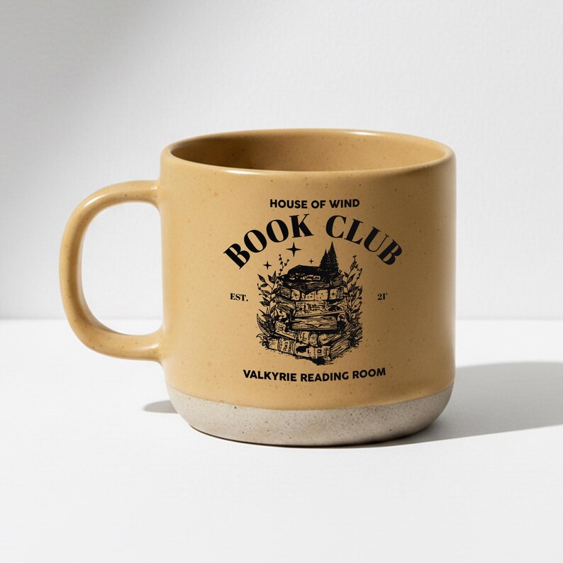 May include: A tan ceramic mug with a white base and a curved handle. The mug features black text that reads "House of Wind Book Club" with an illustration of stacked books. The text "Valkyrie Reading Room" is also printed on the mug.