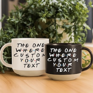 Op de afbeelding: Twee keramische mokken, één crème en één zwart, met de tekst "THE ONE WHERE CUSTOM YOUR TEXT" in een gestileerd lettertype. De mokken hebben een beige basis en een gebogen handvat. De achtergrond toont groen gebladerte.