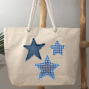 May include: A cream-colored canvas tote bag featuring three star-shaped fabric appliqués. One star is made of blue denim with frayed edges, while the other two stars feature blue and white gingham patterns. The bag has two thick white rope handles.
