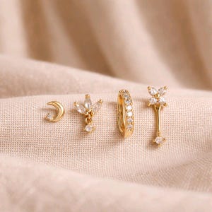 May include: A selection of gold earrings featuring diverse designs. The set includes a crescent moon stud, a floral stud with a dangling gem, a hoop earring with clear stones, and a long floral drop earring with a dangling gem. The earrings are presented on a beige fabric.