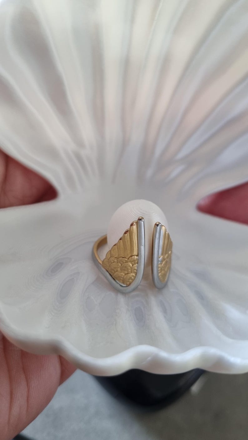 May include: A gold and silver ring with a wing design, set against a white background. The ring features a detailed, textured wing pattern in gold, contrasted by a silver band. The ring is displayed inside a white shell.