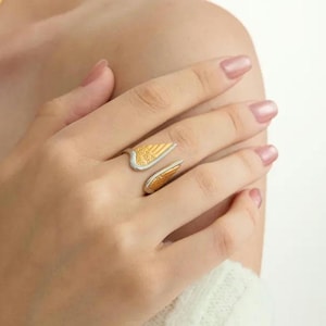 May include: A gold and silver-toned ring in the shape of angel wings. The ring features a textured gold wing design and a silver band. The ring is worn on a finger.