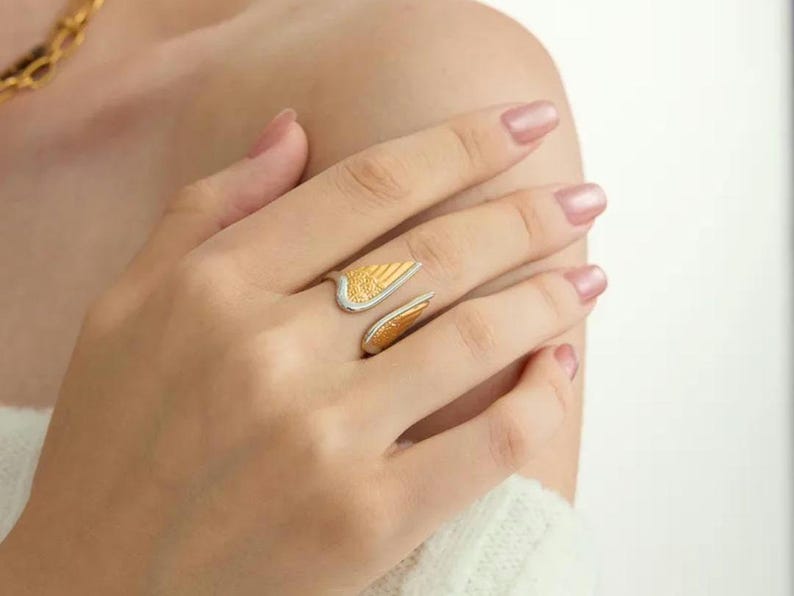 May include: A gold and silver-toned ring in the shape of angel wings. The ring features a textured gold wing design and a silver band. The ring is worn on a finger.
