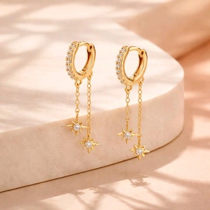Può includere: Orecchini a cerchio dorati con una fila di piccole pietre trasparenti. Ogni orecchino ha una delicata catena con tre ciondoli a forma di stella, ciascuno con una pietra centrale trasparente. Presentati su una superficie chiara.