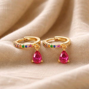 May include: A pair of gold-coloured hoop earrings. The hoops are decorated with small, colourful gemstones. Each earring has a dangling, teardrop-shaped, bright pink gemstone. The earrings are displayed on a beige fabric.