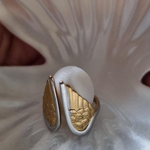 May include: A white pearl ring with gold and silver wing-shaped accents. The ring is set against a white, textured background, creating a striking contrast. The gold wings have a detailed, textured design.