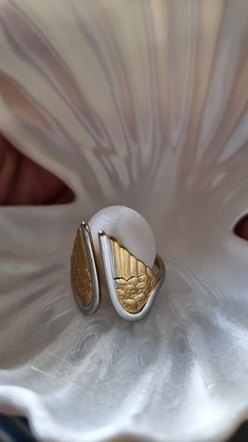 May include: A white pearl ring with gold and silver wing-shaped accents. The ring is set against a white, textured background, creating a striking contrast. The gold wings have a detailed, textured design.