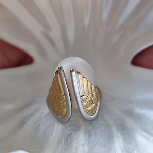 May include: A pair of gold and silver-toned earrings with a wing-like design, resting on a white, textured surface. The earrings feature a detailed floral pattern on the gold portion. The image is well-lit, highlighting the jewelry's intricate details.
