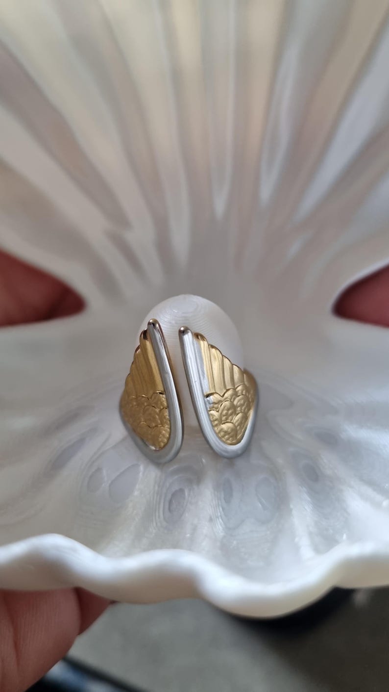 May include: A pair of gold and silver-toned earrings with a wing-like design, resting on a white, textured surface. The earrings feature a detailed floral pattern on the gold portion. The image is well-lit, highlighting the jewelry's intricate details.