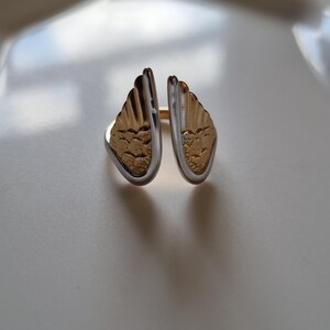 May include: A two-tone ring with a wing design. The ring features gold-colored wings with a textured pattern, framed by a silver-colored band. The ring is set against a white background.