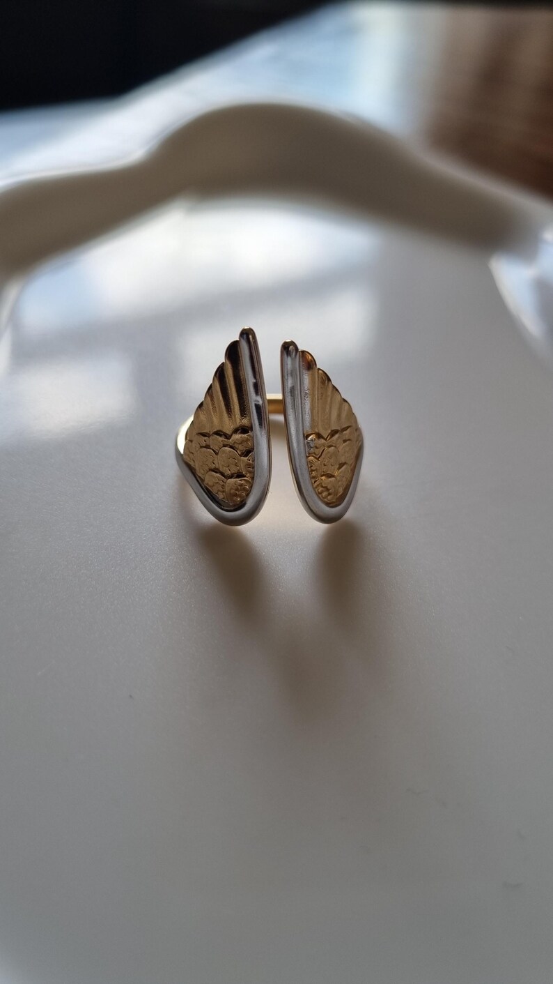 May include: A two-tone ring with a wing design. The ring features gold-colored wings with a textured pattern, framed by a silver-colored band. The ring is set against a white background.