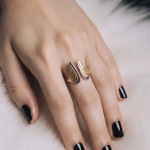 May include: A gold-toned ring with an open design, featuring wing-shaped elements. The ring has intricate detailing and is worn on a hand with black nail polish. The ring's design is reminiscent of angel wings.