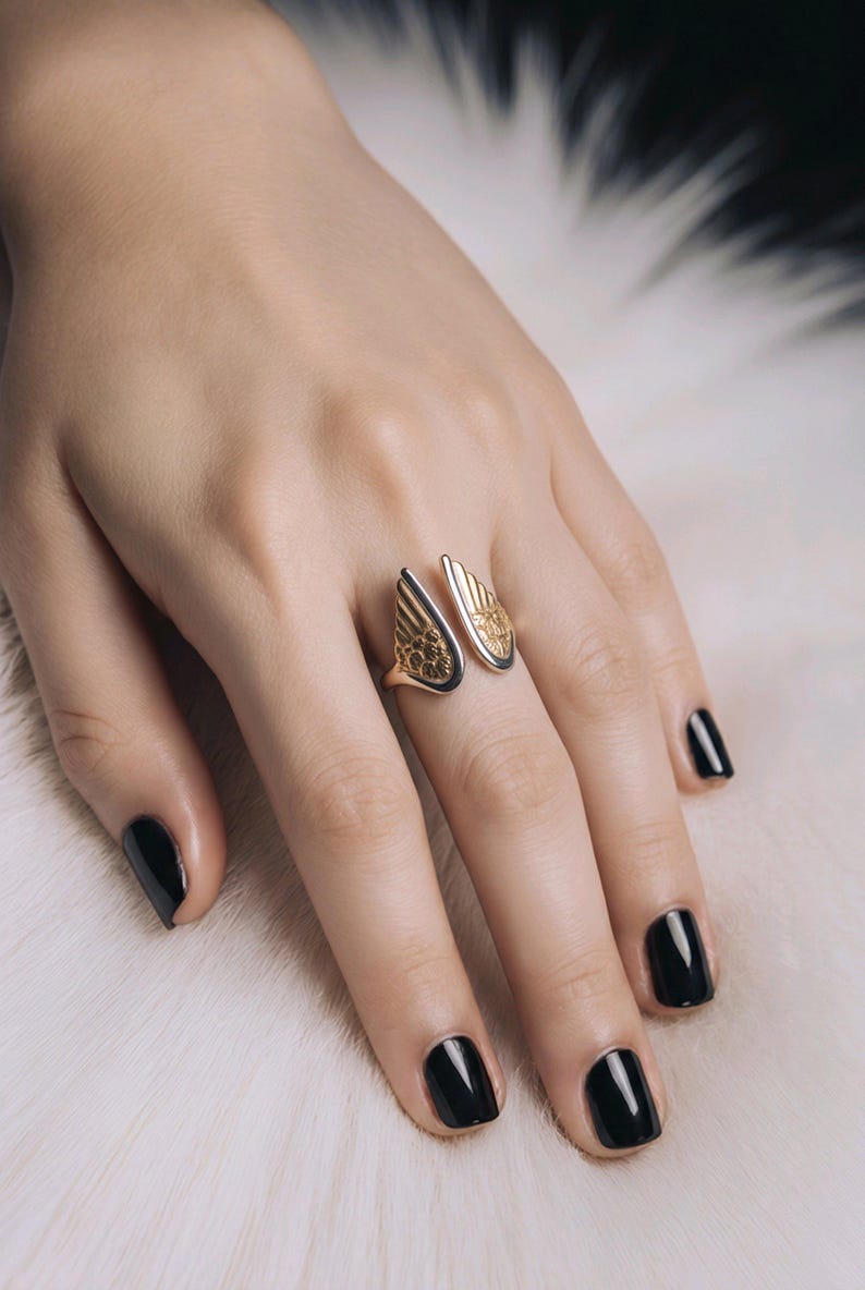 May include: A gold-toned ring with an open design, featuring wing-shaped elements. The ring has intricate detailing and is worn on a hand with black nail polish. The ring's design is reminiscent of angel wings.