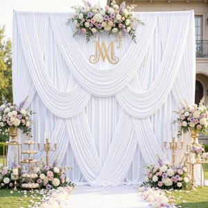 May include: A white draped fabric backdrop with a gold initial "M" and floral arrangements. The scene includes a white aisle, gold candelabras, and additional floral displays, creating an elegant wedding or event setting.