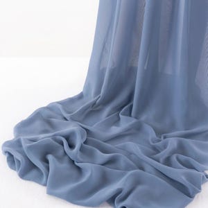 May include: A dusty blue fabric with a sheer, lightweight, and semi-transparent texture. The material is draped in soft, flowing folds that create a series of gentle ripples and curves, showcasing the smooth and consistent matte finish of the textile.