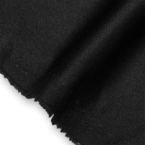 Op de afbeelding: Een stuk zwarte stof met een fijne, uniforme en dichte gebreide textuur. Het materiaal is gevouwen, waardoor zachte, diagonale schaduwen over het oppervlak ontstaan, en heeft een rauwe, licht gerafelde rand aan de linkerkant van het textiel.