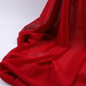 May include: A sheer, vibrant red fabric draped in soft, flowing folds. The material is lightweight and translucent, creating deep shadows and bright highlights within the cascading layers of the textile as it pools and hangs in a vertical orientation.
