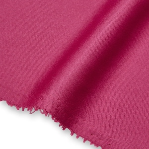 Op de afbeelding: Een stuk magentakleurige stof met een zichtbare geweven textuur en een rauwe, gerafelde rand. Het materiaal is gedrapeerd om zachte plooien te creëren, wat de dieproze kleur en het consistente, strakke weefpatroon over het oppervlak benadrukt.