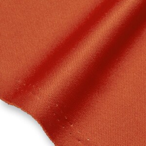 Op de afbeelding: Een close-up van een levendige oranje stof met een fijne, uniforme mesh-achtige textuur. Het materiaal vertoont subtiele plooien en schaduwen die het consistente weefpatroon en de rijke, verzadigde kleur van het textieloppervlak benadrukken.