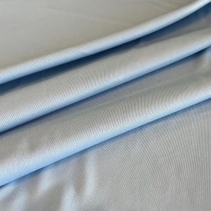 Könnte beinhalten: Ein hellblauer Stoff mit einer feinen, gleichmäßigen Webstruktur. Das Material ist in weichen, diagonalen Falten drapiert, wodurch subtile Schatten und Lichter auf der Oberfläche entstehen, die die glatte, konsistente Farbe und die dichte Webart betonen.