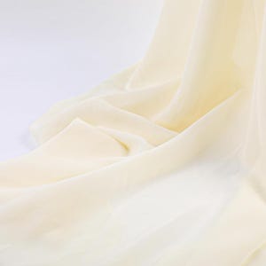 May include: A sheer, lightweight fabric in a soft cream color. The material is draped in loose, flowing folds, creating a series of gentle curves and shadows that highlight the translucent and delicate texture of the textile surface.