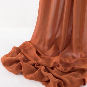 May include: A length of sheer, lightweight fabric in a solid burnt orange color. The material is draped in soft, flowing folds that create a series of gentle curves and shadows, highlighting the translucent quality and fine texture of the textile.
